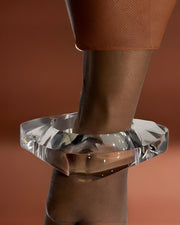 Close-up of the Orbitz Round Lucite Bangle by Nik Spruill on a person's wrist against a brown background. The person is wearing a textured brown sleeve, adding an air of understated luxury.