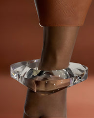 Close-up of the Orbitz Round Lucite Bangle by Nik Spruill on a person's wrist against a brown background. The person is wearing a textured brown sleeve, adding an air of understated luxury.