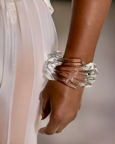 A close-up of a person wearing a unique Ripple Lucite Wide Cuff Bracelet by Nik Spruill, featuring a hand-sculpted liquid wave design on their wrist, next to a light-colored, semi-transparent pleated fabric.