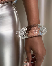 Close-up of a person’s hand wearing a large, clear, hand-sculpted Croc Lucite Cuff Bracelet by Nik Spruill, with a metallic skirt visible in the background.