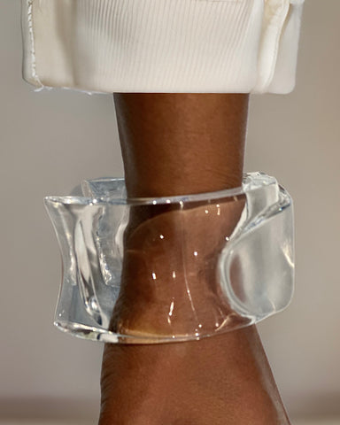A clear, chunky glass or Lucite bracelet called "Tear Drop" by Nik Spruill is worn on a person's wrist, with the end of a white sleeve visible above it.