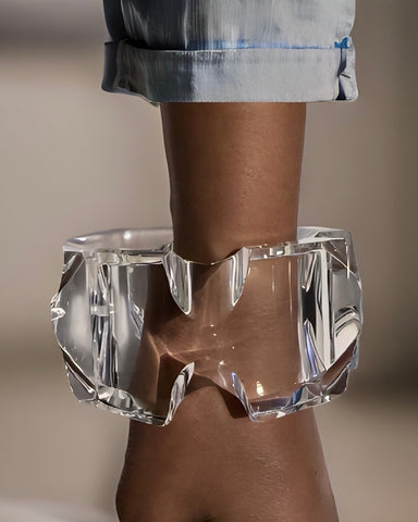 A clear, sculptural Nik Spruill Liquid Lucite Infinity Cuff Bracelet with angular edges adorns an arm wearing a light blue, rolled-up sleeve, showcasing its ultra-modern design.