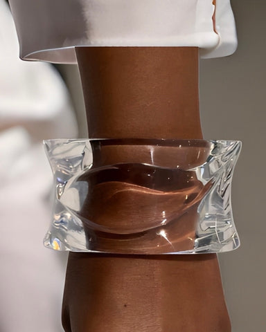 A close-up of a person's arm wearing the large, transparent Luminous Lucite Cuff Bangle Bracelet by Nik Spruill, featuring ripple wave detail, over a white sleeve.