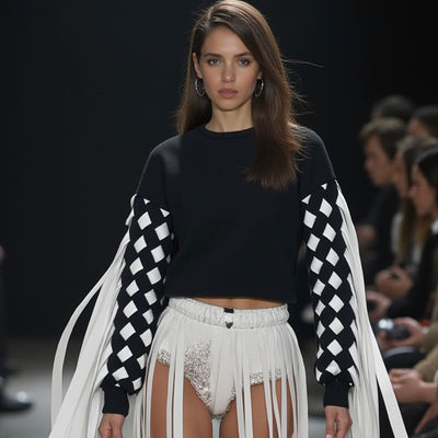 A model walks the runway at the fashion show in a Nik Spruill Stitch Crochet Fringed Top, featuring checkered sleeves with ribbon fringes, paired with white shorts.