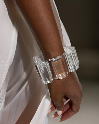 Close-up of a person wearing a hand-sculpted, transparent Geer Lucite Cuff Bracelet by Nik Spruill on their wrist, with sheer white fabric in the background.