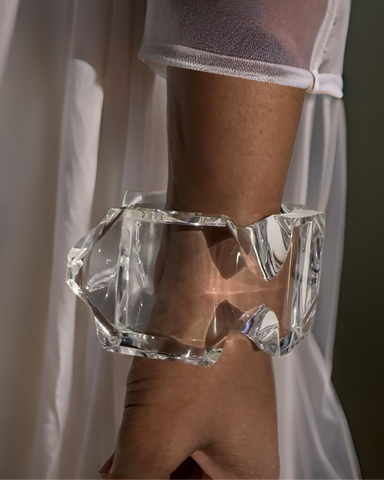 A clear, chunky Nik Spruill Liquid Lucite Infinity Cuff Bracelet shaped like a crystal adorns a person's wrist, seen against the backdrop of sheer, white fabric.