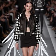 A model wearing a black and white "CHECKMATE FRINGE JACKET" by Nik Spruill, paired with a black skirt and bra top, walks down the runway at a fashion show, with the audience visible in the background.