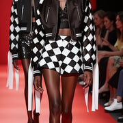 Model on runway in Nik Spruill's CHECKMATE CROCHET SHORT, showcasing black and white color block design with long white ribbons against a vibrant red background. Audience seated behind.