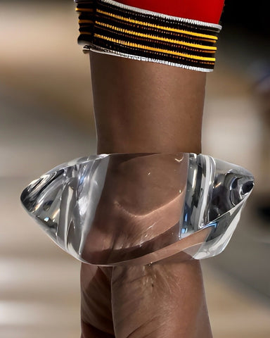 A close-up of the Liquid Lucite Asymmetrical Bangle Bracelet by Nik Spruill on a person's wrist, highlighting the transparency and cut of the hand-sculpted material. The person is wearing a red sleeve with yellow and black stripes.
