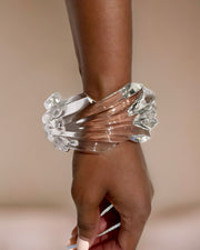 Close-up of a person’s wrist adorned with a transparent, hand-sculpted liquid wave design bracelet on a neutral background—a unique addition boasting the elegance of Nik Spruill's Ripple Lucite Wide Cuff Bracelet.