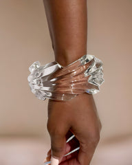 Close-up of a person’s wrist adorned with a transparent, hand-sculpted liquid wave design bracelet on a neutral background—a unique addition boasting the elegance of Nik Spruill's Ripple Lucite Wide Cuff Bracelet.
