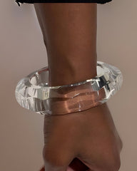 A clear, faceted, cylindrical bracelet worn on a person's wrist, with the hand slightly clenched; the Nik Spruill Bey Stackable Lucite Bangle Bracelet makes for a unique fashion statement.