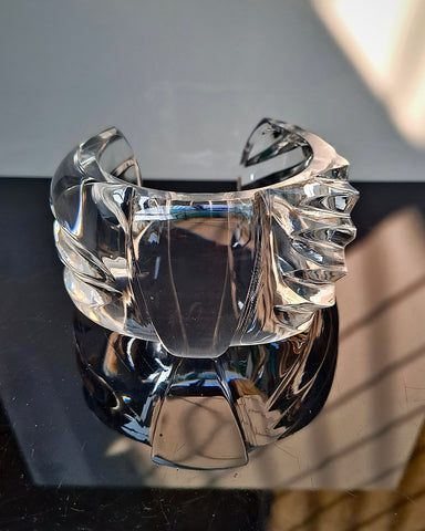 A transparent, intricately designed Bow Lucite Cuff Bracelet by Nik Spruill, resembling a hand-carved lucite creation, sits on a reflective black surface with a white and shadowed background.