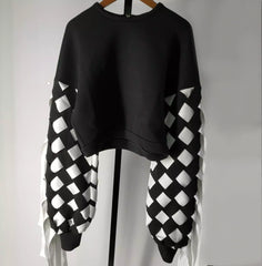 The Stitch Crochet Fringed Top by Nik Spruill is a striking black sweatshirt with unique white and black checkered sleeves, elegantly displayed on a hanger against a plain background, featuring subtle ribbon fringe detailing.