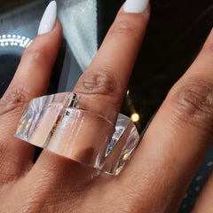 Hand wearing a clear, geometric-shaped ZENUS ring by Nik Spruill on the index finger, with white painted nails and a dark surface in the background.