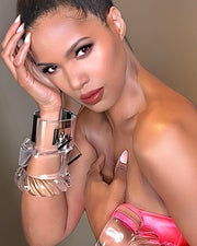 A person with styled hair, wearing a pink strapless top and a Nik Spruill Drop Lock Silver Lucite Bangle Bracelet, poses elegantly with one hand on their face and the other on their chest.