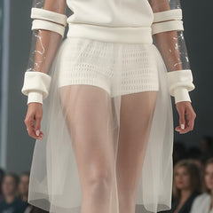 A model on the runway showcases the Nik Spruill HARMONY HALO SHORT, a white outfit featuring a sheer skirt and transparent sleeves with elegant laser-cut neoprene details.