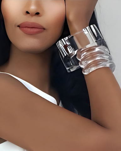 A person with long dark hair and smooth skin, wearing a white top, showcases two large hand-carved Lucite ICEBERG bracelets by Nik Spruill on their wrist.