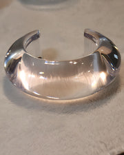 The ARCTIC by Nik Spruill, a hand-polished clear acrylic cuff bracelet, rests on a light-colored surface, showcasing its elegant curves and refined simplicity.