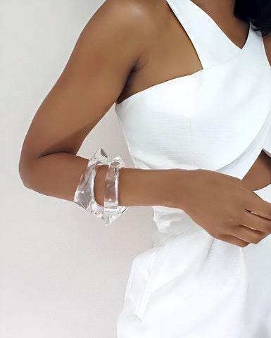 Close-up of a person wearing "ICICLE," a sleeveless white dress with cut-out details by Nik Spruill, paired with two lucite bracelets on their left wrist against a plain background.