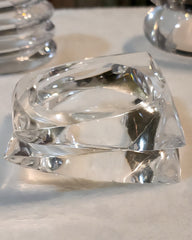 A close-up of two stacked, ICICLE hand-carved, transparent glass bracelets by Nik Spruill, placed on a white surface.