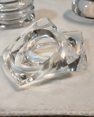 Two clear, intertwined ICICLE glass rings from Nik Spruill rest on a soft white surface, reflecting light. A hand-carved bangle glimmers alongside them, with other glass objects visible in the blurred background.