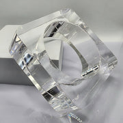 The OHSO by Nik Spruill is a square-shaped, clear Lucite object featuring a round hole in the center, elegantly positioned on its edge against a gray and white background, exuding the sophistication of a hand-carved piece.