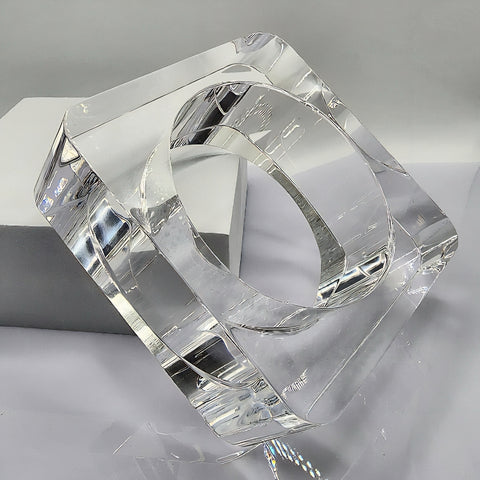The OHSO by Nik Spruill is a square-shaped, clear Lucite object featuring a round hole in the center, elegantly positioned on its edge against a gray and white background, exuding the sophistication of a hand-carved piece.