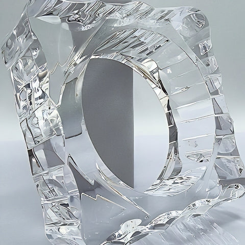 The METROPOLIS by Nik Spruill is a clear, intricately designed glass sculpture, reminiscent of a hand-polished Lucite piece, featuring a central circular hole and various angular patterns. It is displayed on a minimalist light-colored backdrop.