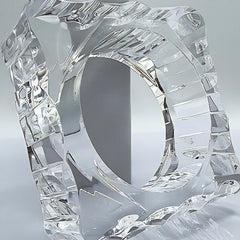 The METROPOLIS by Nik Spruill is a clear, intricately designed glass sculpture, reminiscent of a hand-polished Lucite piece, featuring a central circular hole and various angular patterns. It is displayed on a minimalist light-colored backdrop.