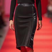 A model showcases the FREEDOM MIDI SKIRT by Nik Spruill, featuring a fitted black design with eel skin leather and shiny side panels, paired with a black top.