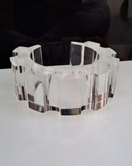 A clear, multi-faceted acrylic sculpture with a wavy, geometric design sits on a white surface against a dark background, reminiscent of the modern design seen in the Nik Spruill Geer Lucite Cuff Bracelet.