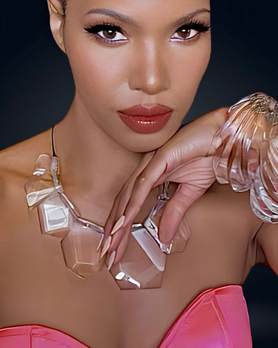 A person with a neutral facial expression, wearing a pink top, the Nik Spruill Tribal Liquid Lucite Sculptural Necklace that exudes modern luxury, and a matching clear bracelet against a dark background.