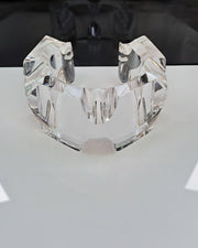 A clear, intricately-cut Liquid Lucite Infinity Cuff Bracelet by Nik Spruill is displayed on a reflective surface.