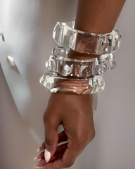 A hand with light brown skin is shown wearing the Nik Spruill Hive Stackable Lucite Bangle Bracelets, featuring four large, clear geometric shapes. The unique design beautifully complements the person's long, pointed white nail polish, making it perfect to elevate your style.