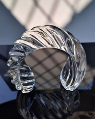 The Wiggle Lucite Cuff Bracelet by Nik Spruill is showcased on a reflective surface, featuring its clear, twisted design with a geometric shadow pattern in the background.