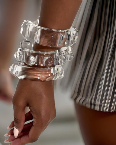 Close-up of a person wearing several large Liquid Lucite Raindrop Cuff Bangles by Nik Spruill on their wrist, exuding elegance and luxury, with a striped fabric in the background.