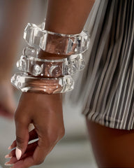 Close-up of a person wearing several large Liquid Lucite Raindrop Cuff Bangles by Nik Spruill on their wrist, exuding elegance and luxury, with a striped fabric in the background.