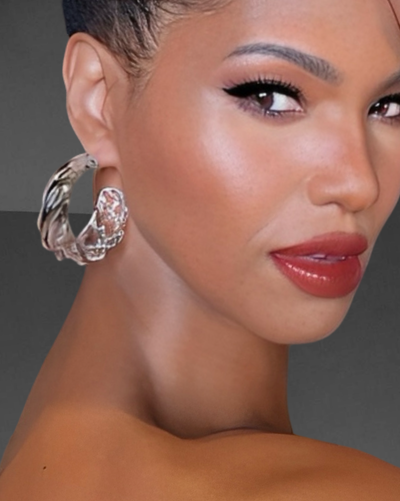 Close-up of a person looking over their shoulder. They have short hair, bold makeup, and wear large, modern Liquid Lucite Wave Hoop Earrings by Nik Spruill. The background is plain gray.