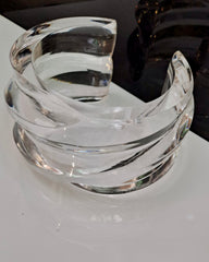 A clear, hand-sculpted Nik Spruill Liquid Lucite Wave Cuff Bracelet with a twisted design, resting on a reflective white surface.