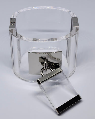 Introducing the "Drop Lock Silver Lucite Bangle Bracelet" by Nik Spruill, a meticulously hand-carved lucite cuff that showcases a sleek rectangular silver clasp with a magnetic closure, elegantly adorned with a metallic ring and rectangular charm.