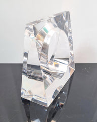 Displayed on a reflective, dark surface, the Liquid Lucite Wedge Bangle Bracelet by Nik Spruill features a clear, geometric design with a triangular shape and circular cutout, reminiscent of sophisticated jewelry.