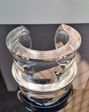 The Dream Rain Lucite Cuff Bracelet by Nik Spruill is exhibited on a clear, curved acrylic display stand with a reflective black surface, designed to hold and showcase items with timeless sophistication.