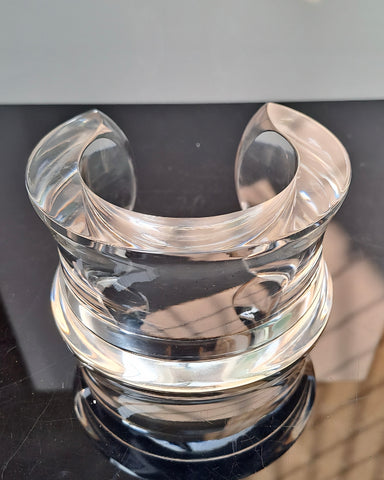 The Dream Rain Lucite Cuff Bracelet by Nik Spruill is exhibited on a clear, curved acrylic display stand with a reflective black surface, designed to hold and showcase items with timeless sophistication.
