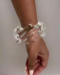 A hand with light pink nails wears a thick, clear Spiral Liquid Lucite Cuff Bracelet by Nik Spruill, featuring cascading wave details against a neutral background.