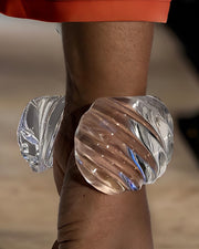 Close-up of the Wiggle Lucite Cuff Bracelet by Nik Spruill on a model's wrist, with part of an orange sleeve visible in the background. The handcrafted bracelet features a spiral design and is translucent.