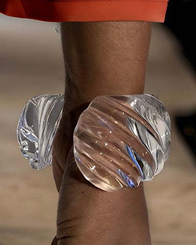 Close-up of the Wiggle Lucite Cuff Bracelet by Nik Spruill on a model's wrist, with part of an orange sleeve visible in the background. The handcrafted bracelet features a spiral design and is translucent.