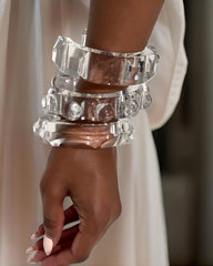 A person wearing multiple large, clear, geometric-shaped bracelets on their wrist, including a sophisticated Nik Spruill Link Lucite Unisex Tennis Cuff Bracelet. The background is blurred and the person is dressed in a light-colored, flowing garment.