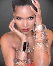 A person with a serious expression, wearing large, clear Nik Spruill Liquid Lucite Dome Cuff Bracelets on both wrists, poses with hands on face and head against a neutral background.