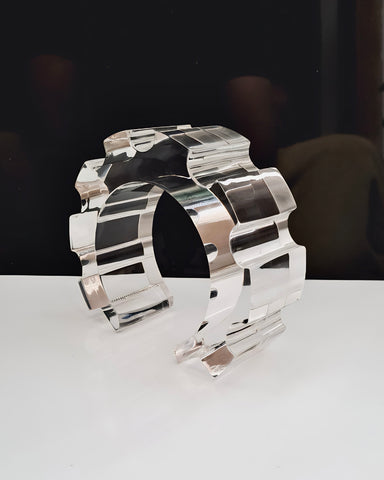 The Geer Lucite Cuff Bracelet by Nik Spruill is displayed on a white surface with a black backdrop. This hand-sculpted piece, crafted from transparent material, features a segmented, geometric arch-like design that epitomizes modern aesthetics.